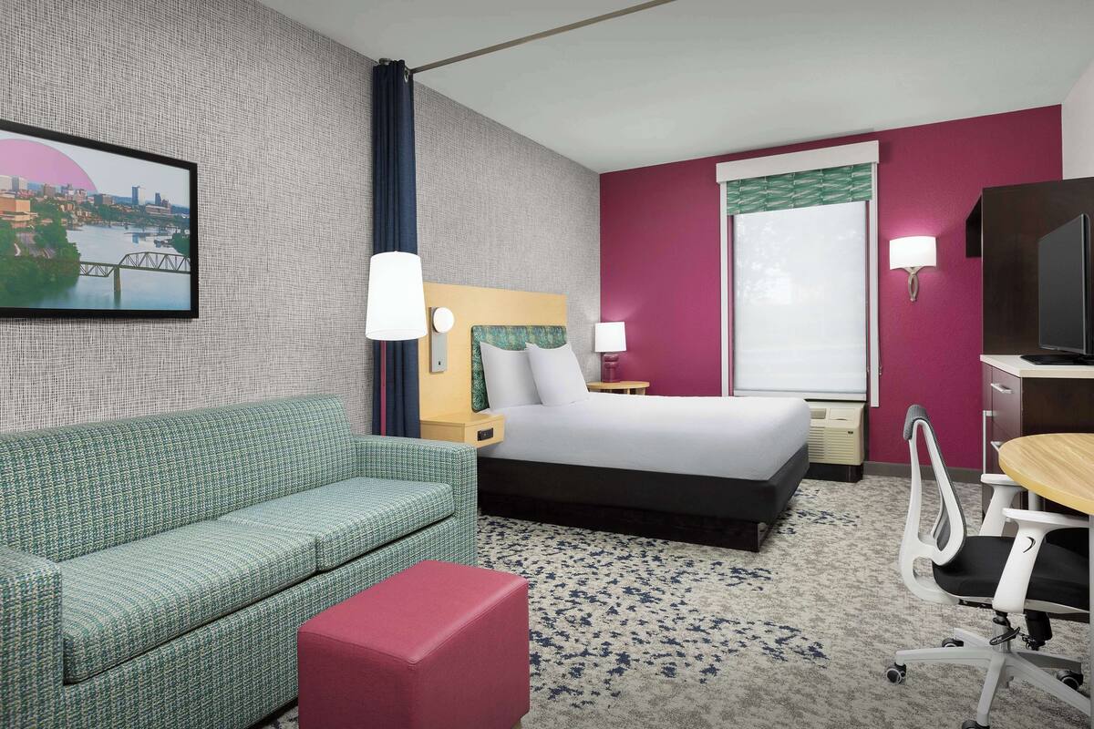 Une chambre d’hôtel avec un lit, un canapé vert, un pouf rouge, un bureau avec une chaise et un téléviseur.