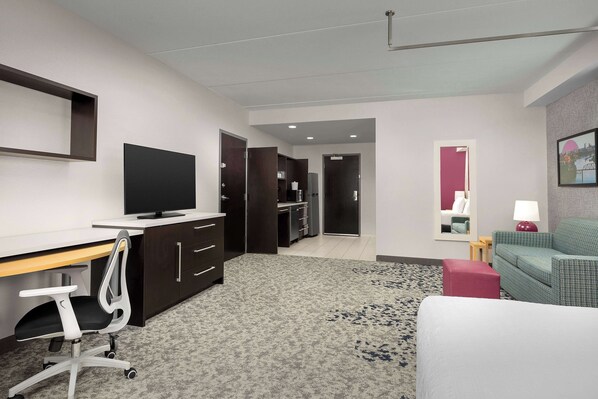 Habitación de hotel con escritorio, televisor, sofá, reposapiés rojo y alfombra estampada.