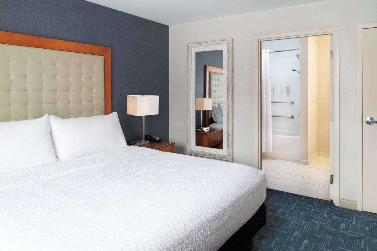 Een hotelkamer met een groot bed, een nachtkastje met lamp, een spiegel en een deur naar de badkamer.