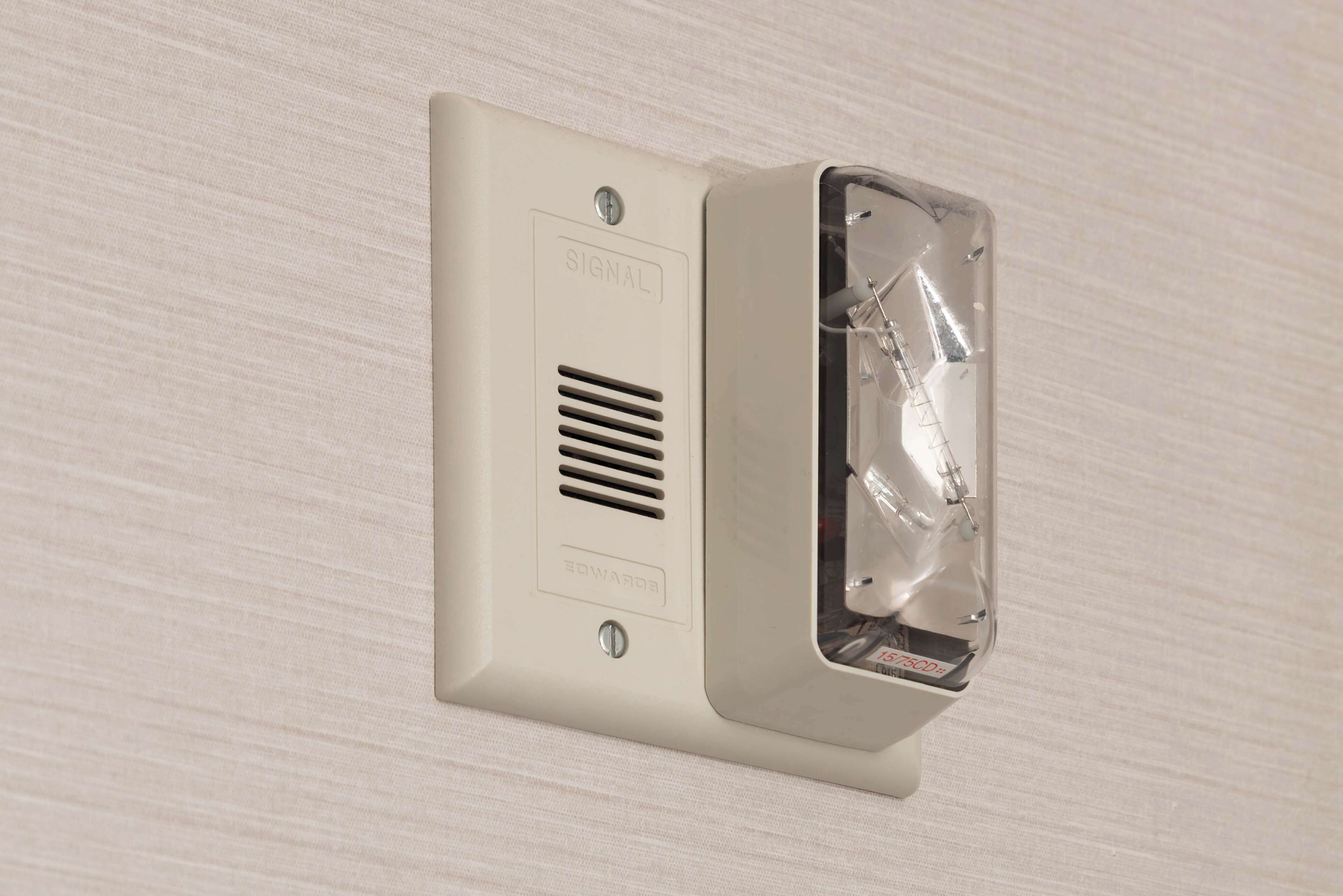 Detector de fumaça fixado na parede, com janela de vidro transparente e revestimento branco.
