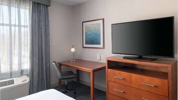 A hotel room with a bed, a desk with a chair, a television on a wooden cabinet, a framed picture on the wall, and a window with curtains.