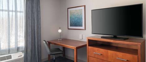 A hotel room with a bed, a desk with a chair, a television on a wooden cabinet, a framed picture on the wall, and a window with curtains.