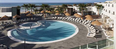 A pool with sun loungers and umbrellas, a beach view, and a modern building.