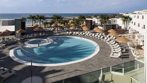 En pool med solstolar och parasoller, utsikt över stranden och en modern byggnad.