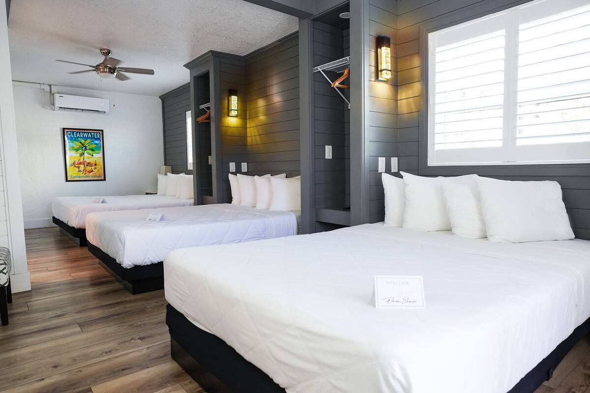 Habitación de hotel con tres camas, un ventilador de techo y un ventanal con persianas.