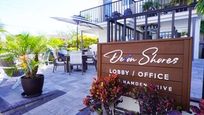 Un letrero de madera con la inscripción 'Devon Shares Lobby/Office' y 'HAMDEN DRIVE', rodeado de plantas en macetas y mobiliario exterior.