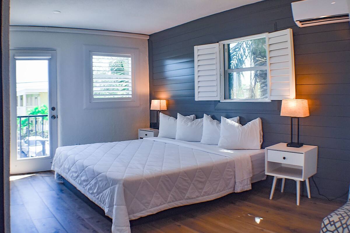 Un dormitorio con cama, dos mesitas de noche, una ventana con contraventanas y un equipo de aire acondicionado.