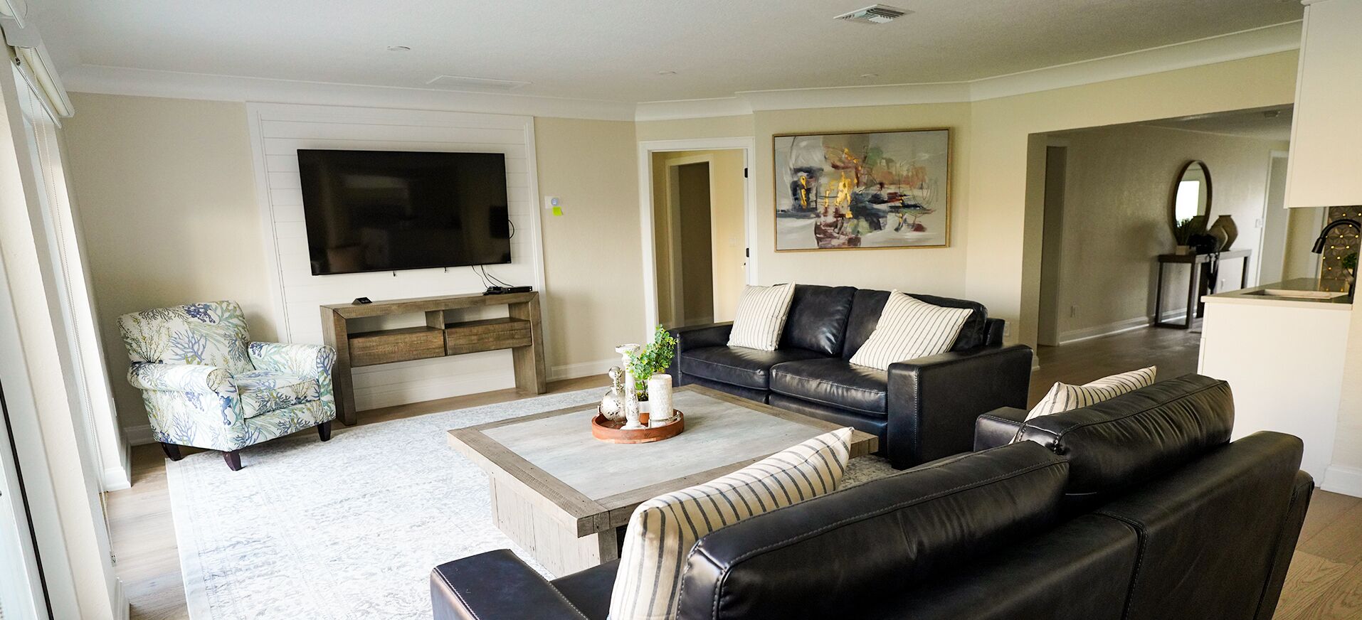 Una sala de estar con un televisor de pantalla plana grande, un sofá de cuero negro, un sillón estampado y una mesa de centro con objetos decorativos.