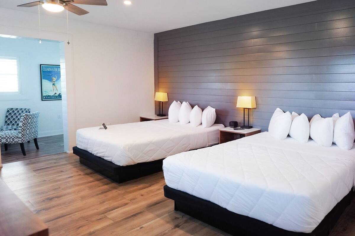 Habitación de hotel con dos camas, suelo de madera, ventilador de techo e iluminación instalada en la pared.