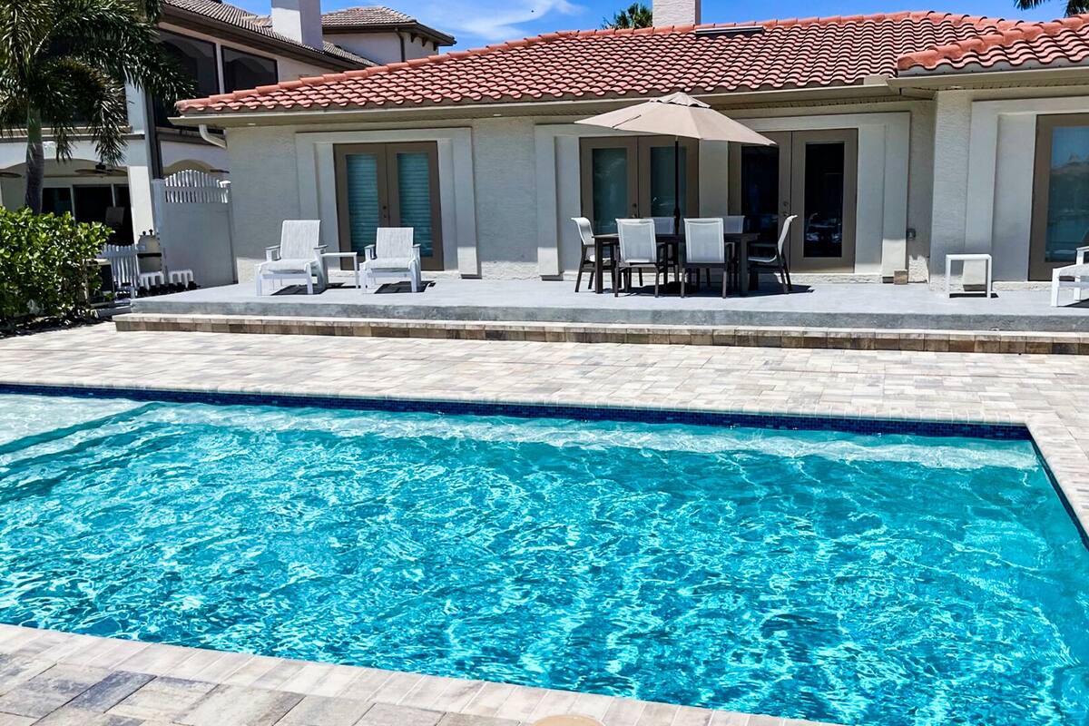 Una piscina con solárium azulejado, mobiliario exterior y un edificio con tejado rojo.
