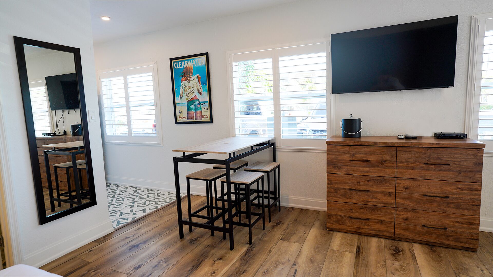 A modern kitchen with a wooden floor, a dining table with stools, a TV mounted on the wall, and a window with blinds.