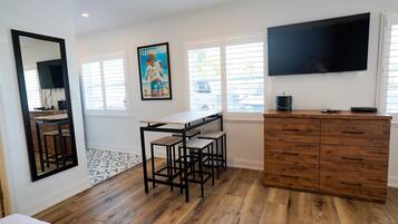 A modern kitchen with a wooden floor, a dining table with stools, a TV mounted on the wall, and a window with blinds.