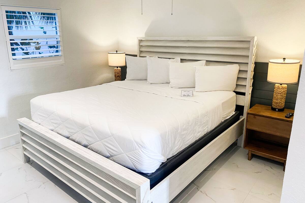 Una cama bien hecha con un cabecero, dos mesitas de noche con lámparas, y un cartel sobre la cama.