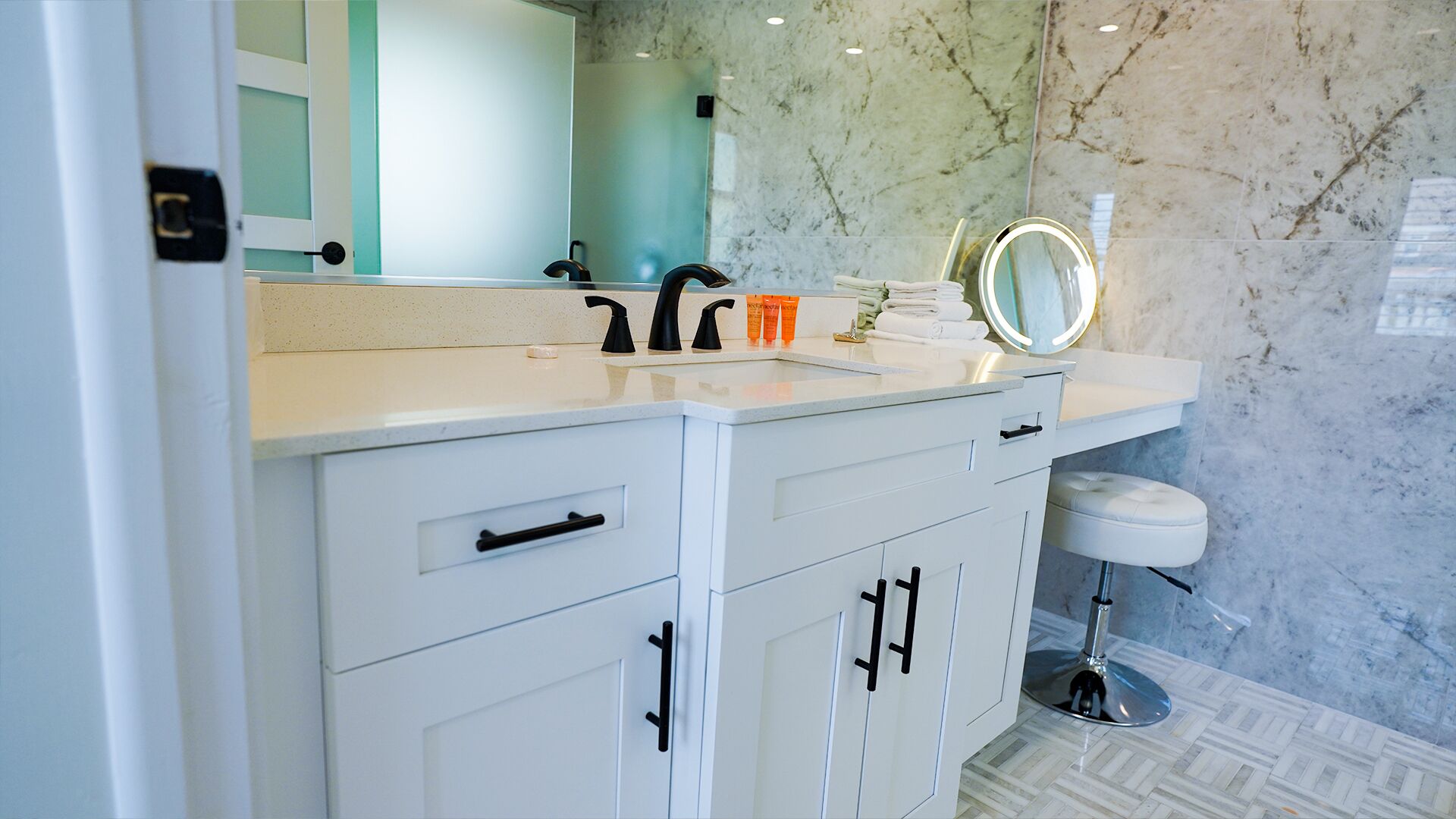 Un baño con tocador blanco, lavamanos, espejo con luces y pared de mármol.
