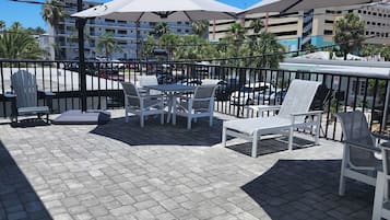 An outdoor patio with white furniture and umbrellas, surrounded by a black metal fence, with buildings and palm trees in the background.