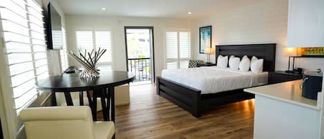 A modern bedroom with a large bed, a desk, and a chair. There is a balcony with a view of the outdoors.