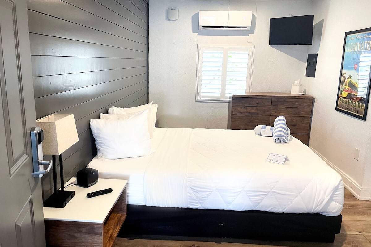 Un dormitorio con cama, mesita de noche, escritorio, cómoda, ventilador de techo, aire acondicionado, ventana e imagen enmarcada en la pared.