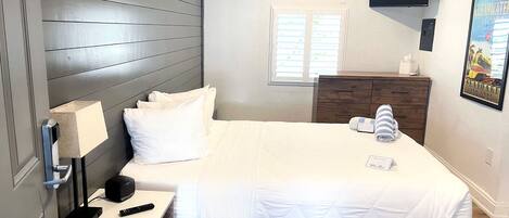 A bedroom with a bed, a nightstand, a desk, a dresser, a ceiling fan, an air conditioning unit, a window, and a framed picture on the wall.
