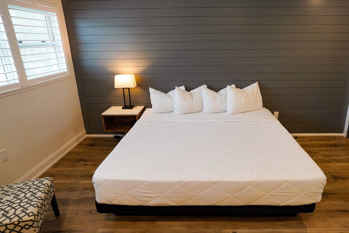 Una cama bien hecha con edredón blanco acolchado, una lámpara de noche y una mesita de madera.