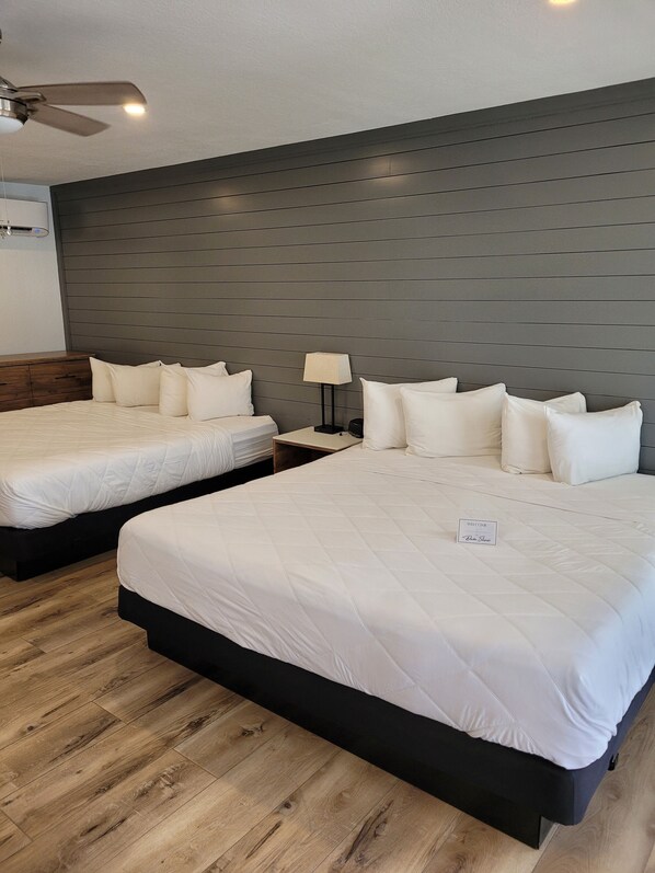 Habitación de hotel con dos camas, piso de madera, cabecera oscura, ventilador de techo y mesita de noche con lámpara.