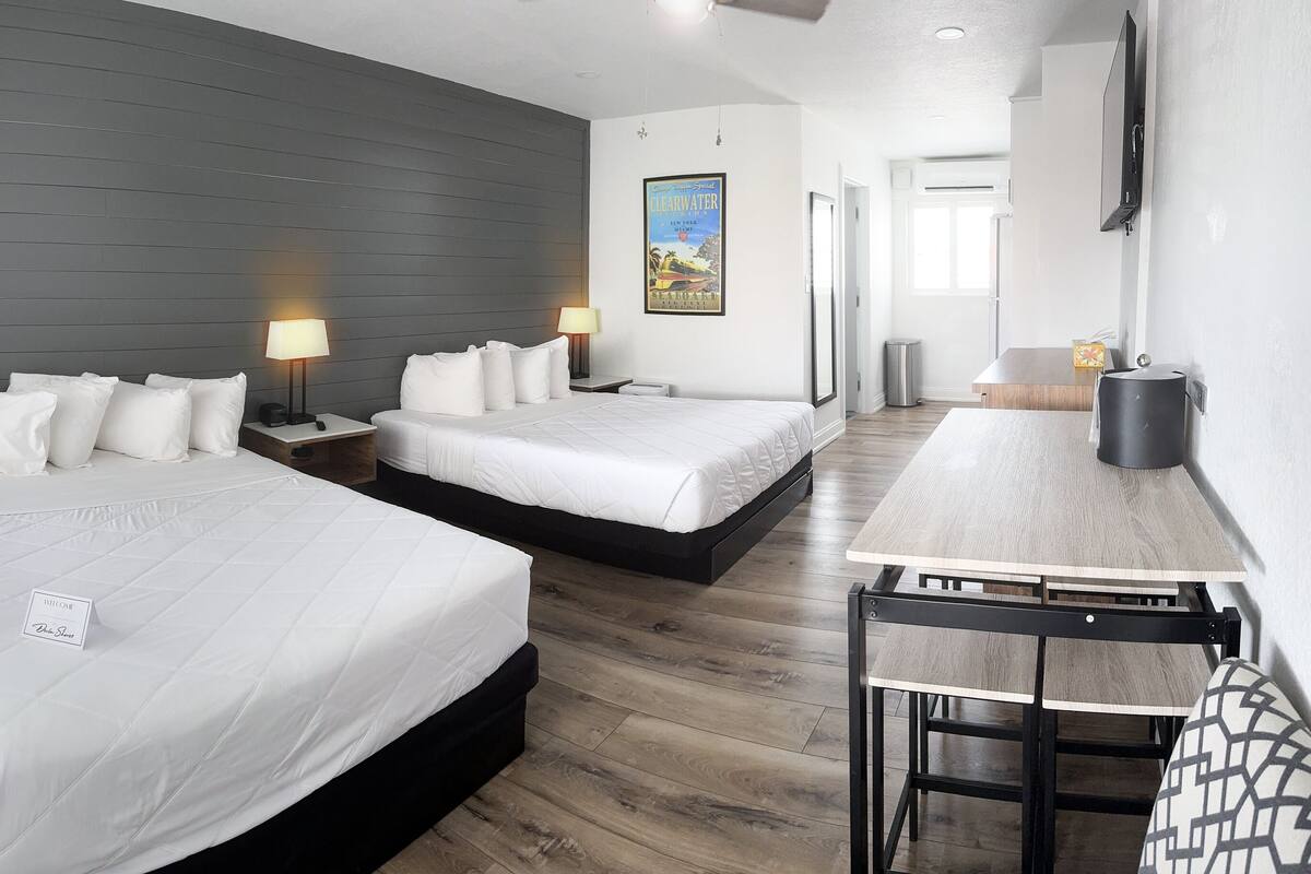Habitación de hotel con dos camas, un escritorio y una silla. Se observan mesitas de noche con lámparas, un cartel en la pared y vistas al exterior a través del ventanal.