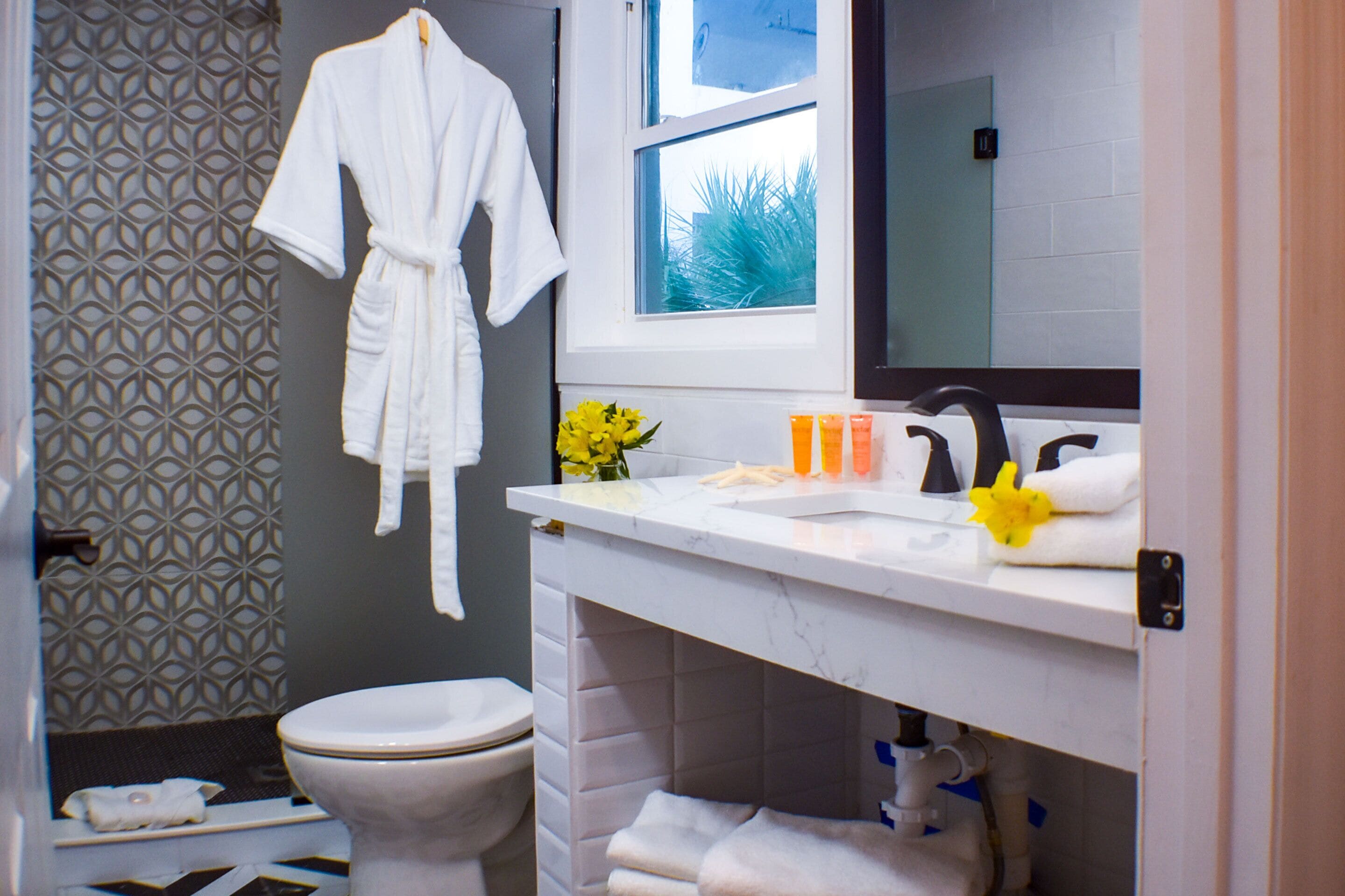 A bathroom with a toilet, a white robe hanging, a sink with toiletries, and a window with a view.