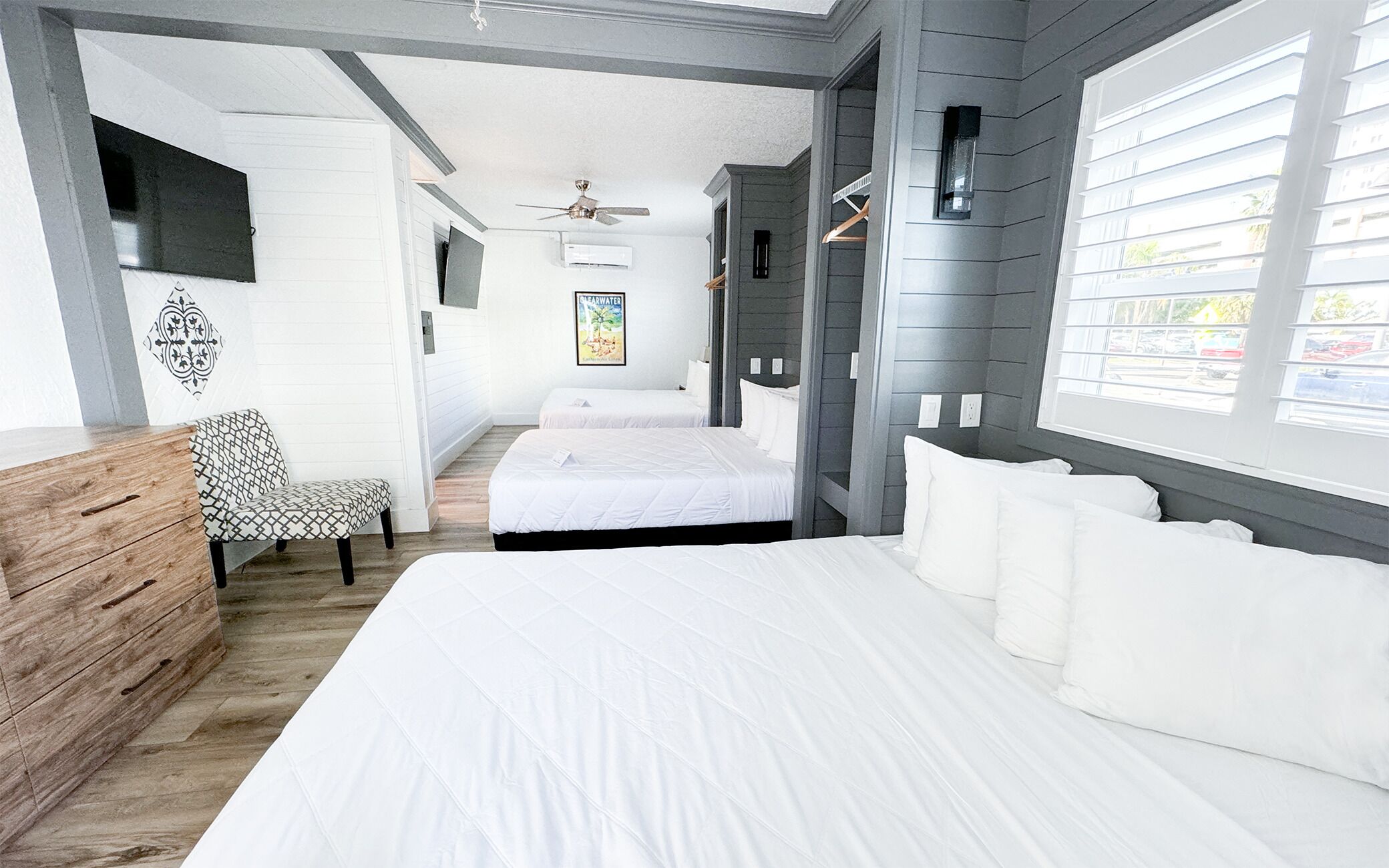 Ein Hotelzimmer mit einem großen Bett, zwei kleineren Betten, einer Holzkommode, einer Bank und einem Deckenventilator.