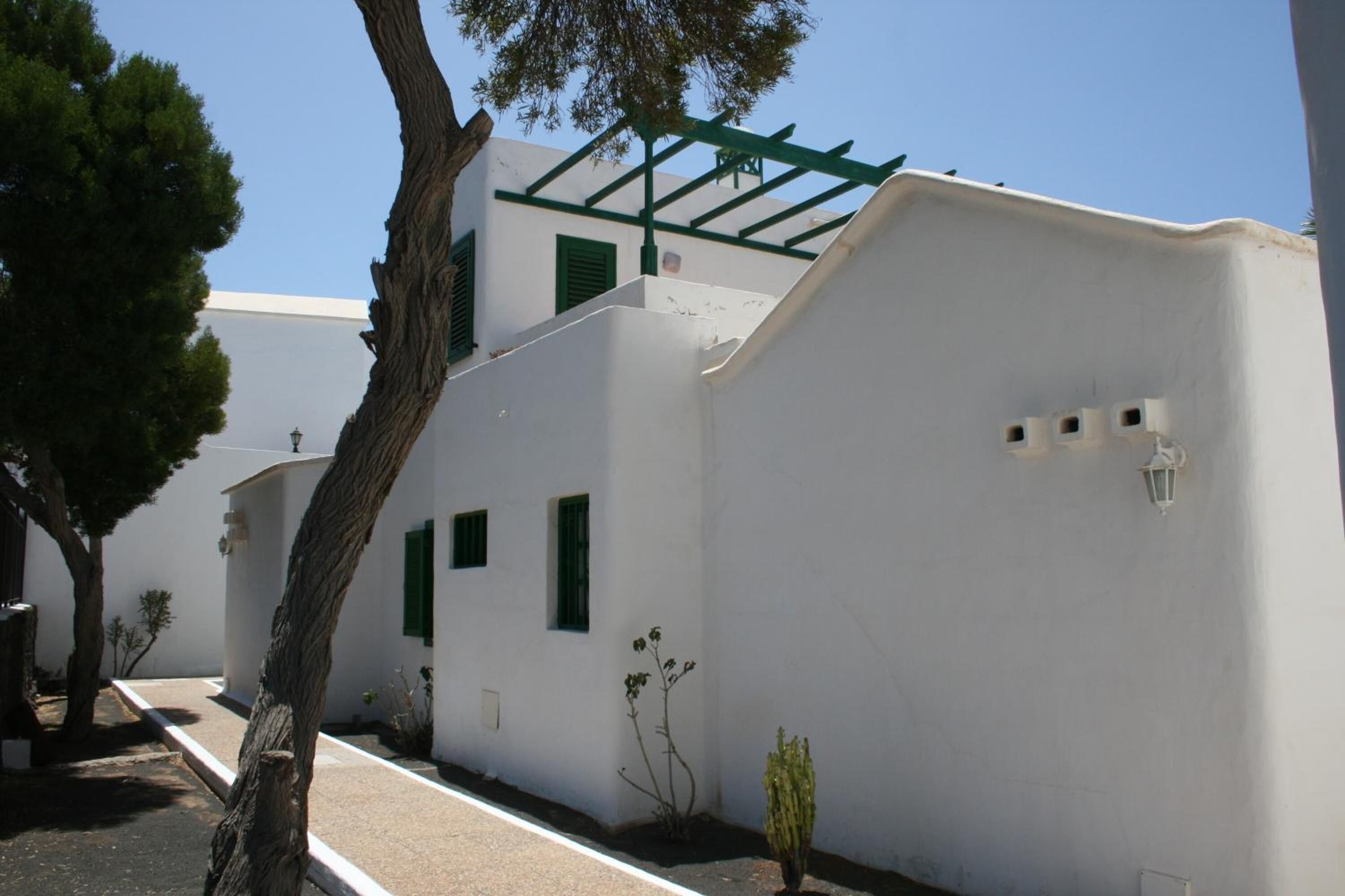 Un edificio blanco con contraventanas verdes, un árbol en primer plano y un cielo despejado.
