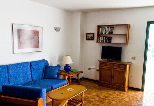 A living room with a blue sofa, a small table with a lamp, a wooden cabinet, and a television. - Apartamentos las Acacias (Tías)