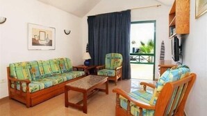 Uma sala de estar com sofá, poltrona e mesa de centro. Há uma janela com cortinas, um quadro na parede e uma televisão sobre um suporte de madeira.