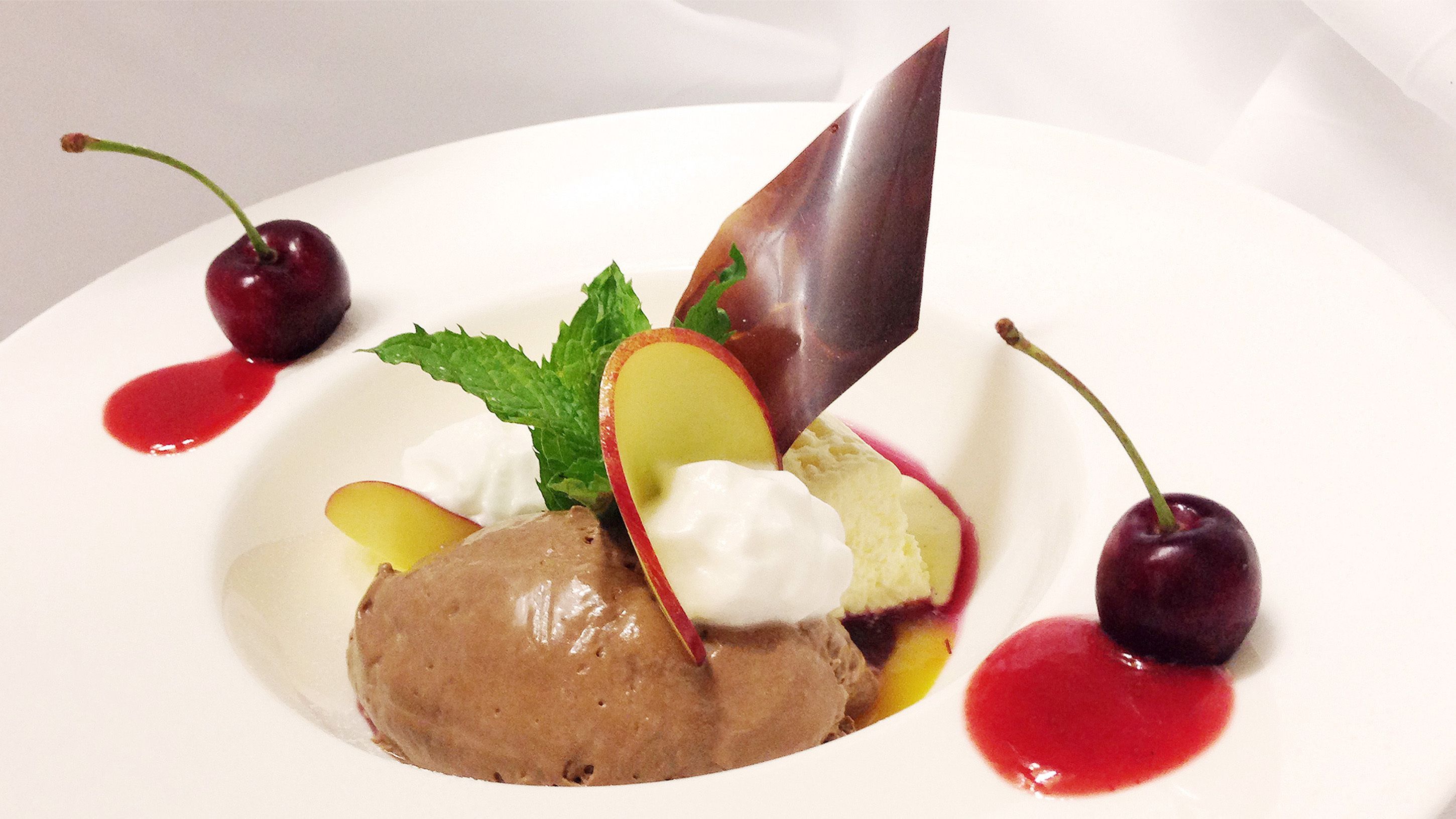 A dessert plate with a scoop of ice cream, a piece of chocolate, cherries, and a mint leaf.