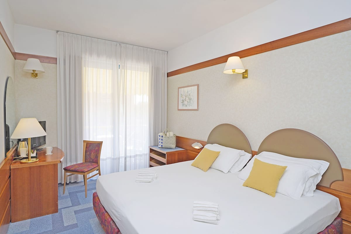 A hotel room with a bed, a desk with a lamp, a chair, and a nightstand with a lamp.