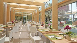 An outdoor dining area with wooden tables and white wicker chairs, surrounded by glass walls and large windows. - Hotel Sportsman (Bardolino)