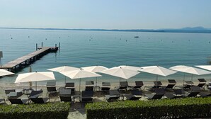 A beach with lounge chairs and umbrellas, a pier extending into calm waters, and distant mountains. - Hotel Sportsman (Bardolino)