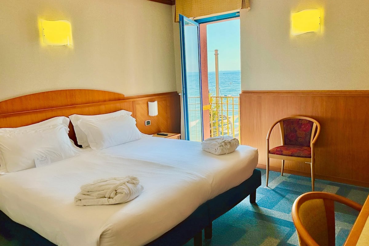 A hotel room with a large bed, a chair, and a view of the sea through an open door.