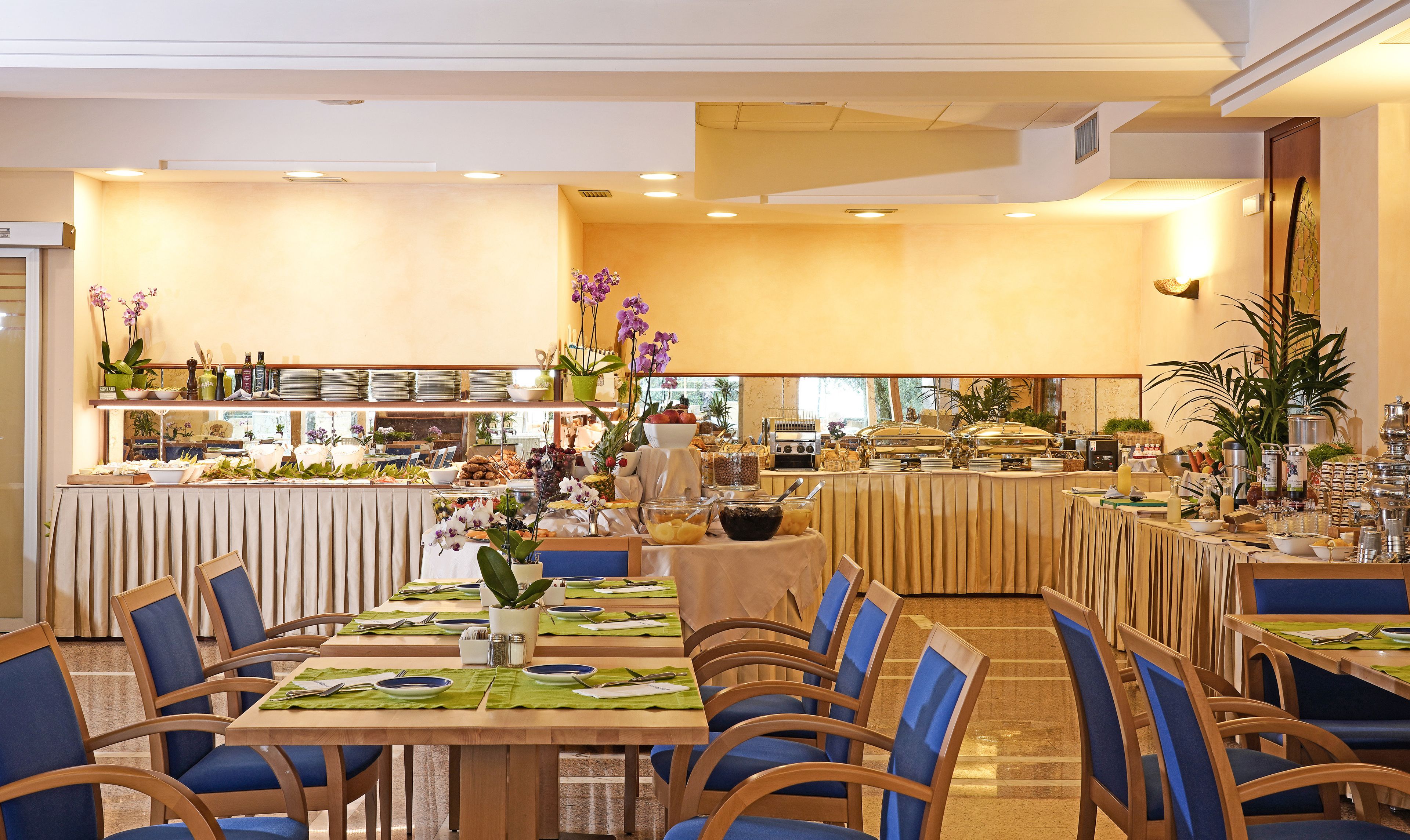 Un restaurante bien iluminado con varias mesas de comedor dispuestas con sillas azules, una zona de bufé con diversos alimentos y una decoración interior cálida.