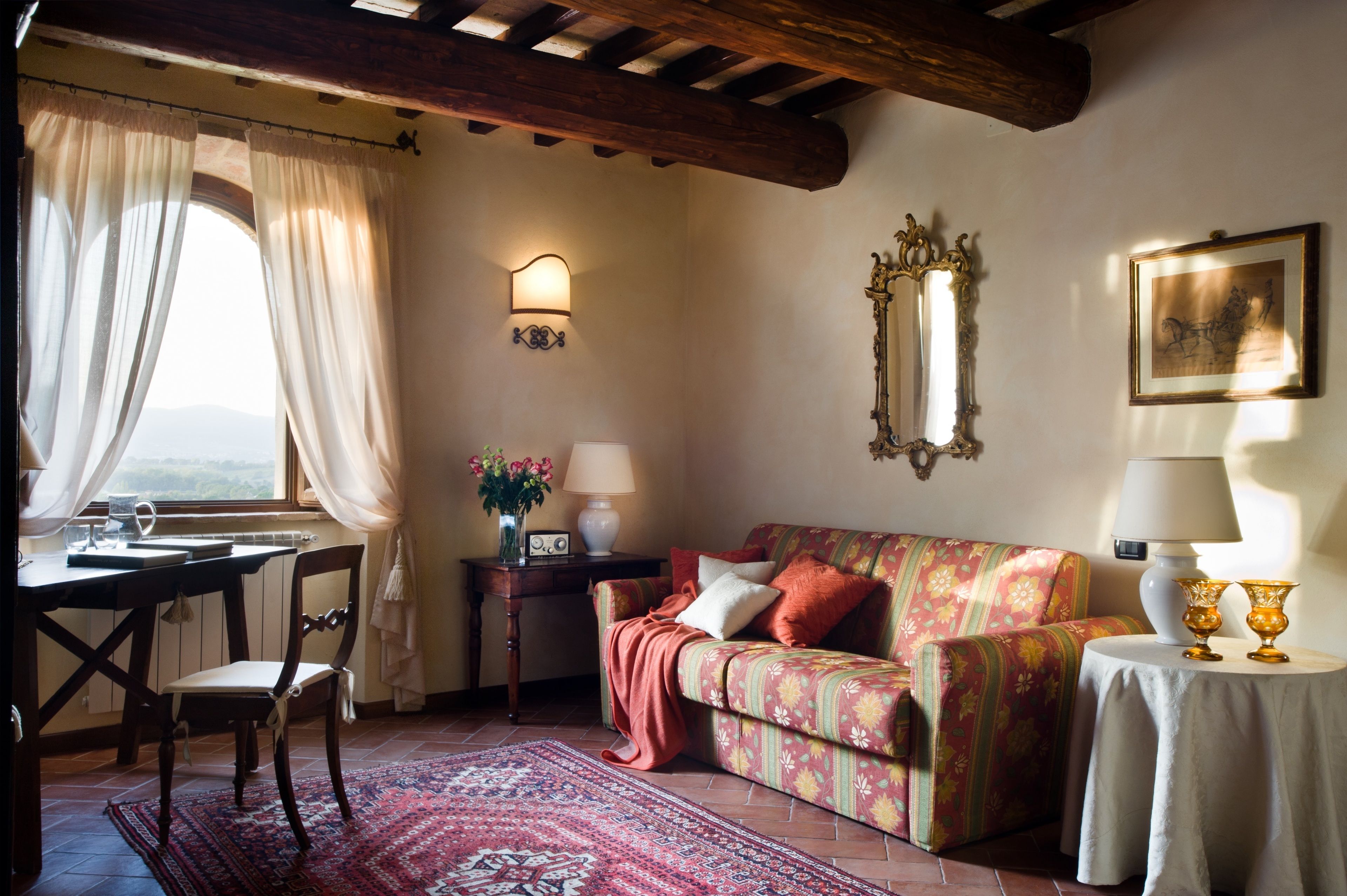 Ein gemütliches Zimmer mit Holzbalkendecke, einer blumengedeckten Couch, einem Tisch mit Lampe und einem Fenster mit Vorhängen.