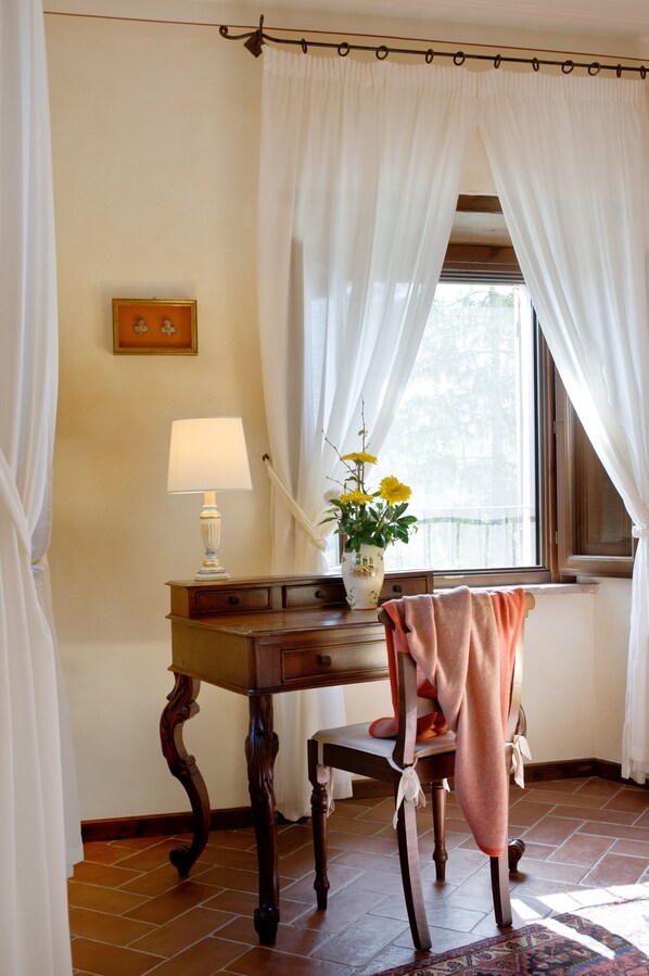 Una habitación con una mesa de madera, una silla, una lámpara, un florero con flores y una ventana con cortinas.
