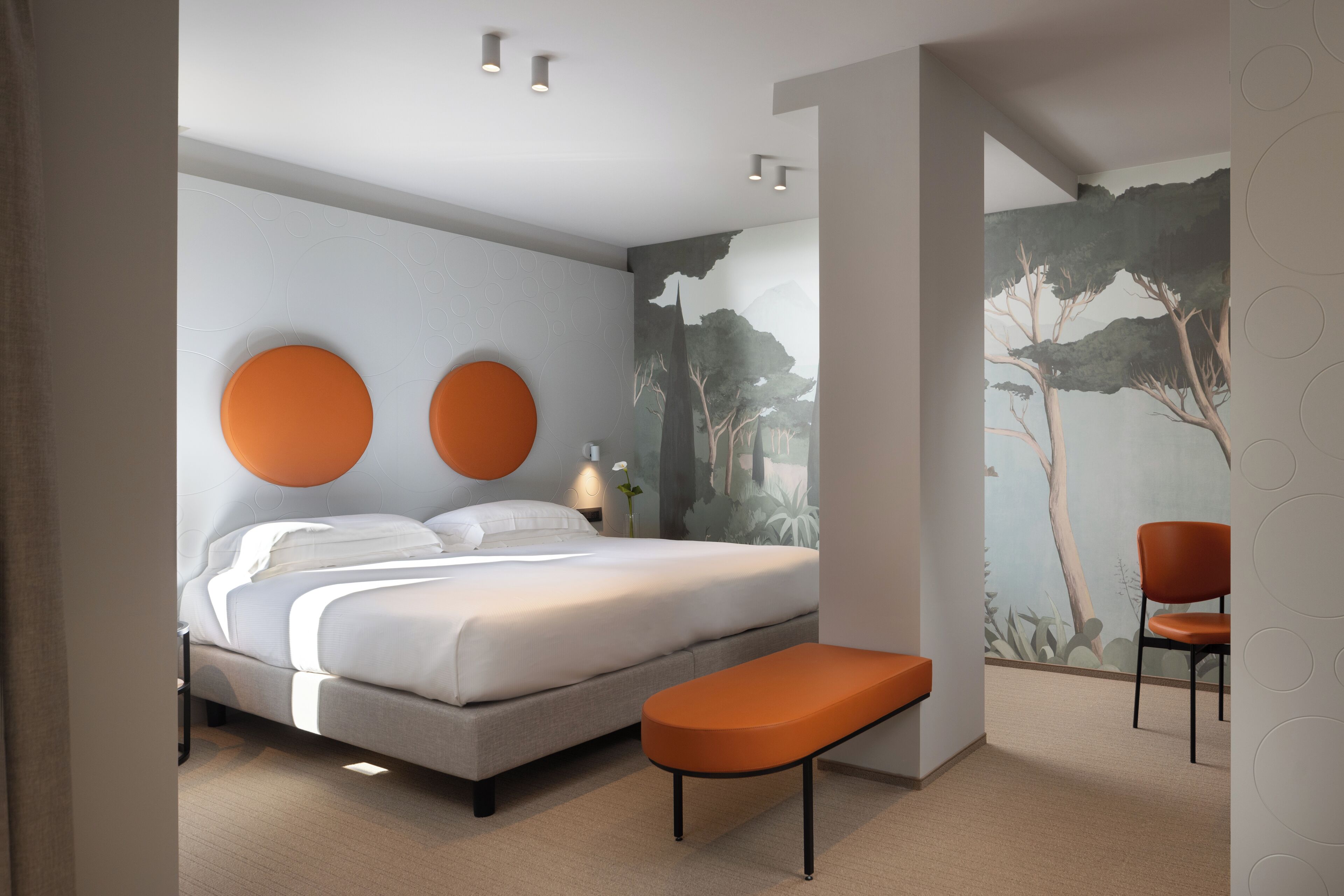 A modern bedroom with a large bed, two wall-mounted circular light fixtures, a bench, and a mural of trees.