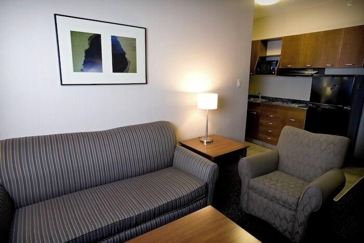 Un espacio de estar con un sofá, un sillón y una mesita. Hay una lámpara en una mesita auxiliar y un cuadro enmarcado en la pared.
