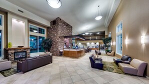 A spacious lobby with a stone fireplace, a long leather sofa, a reception desk, and a view of the kitchen area. - Heritage Inn & Suites (Brooks)