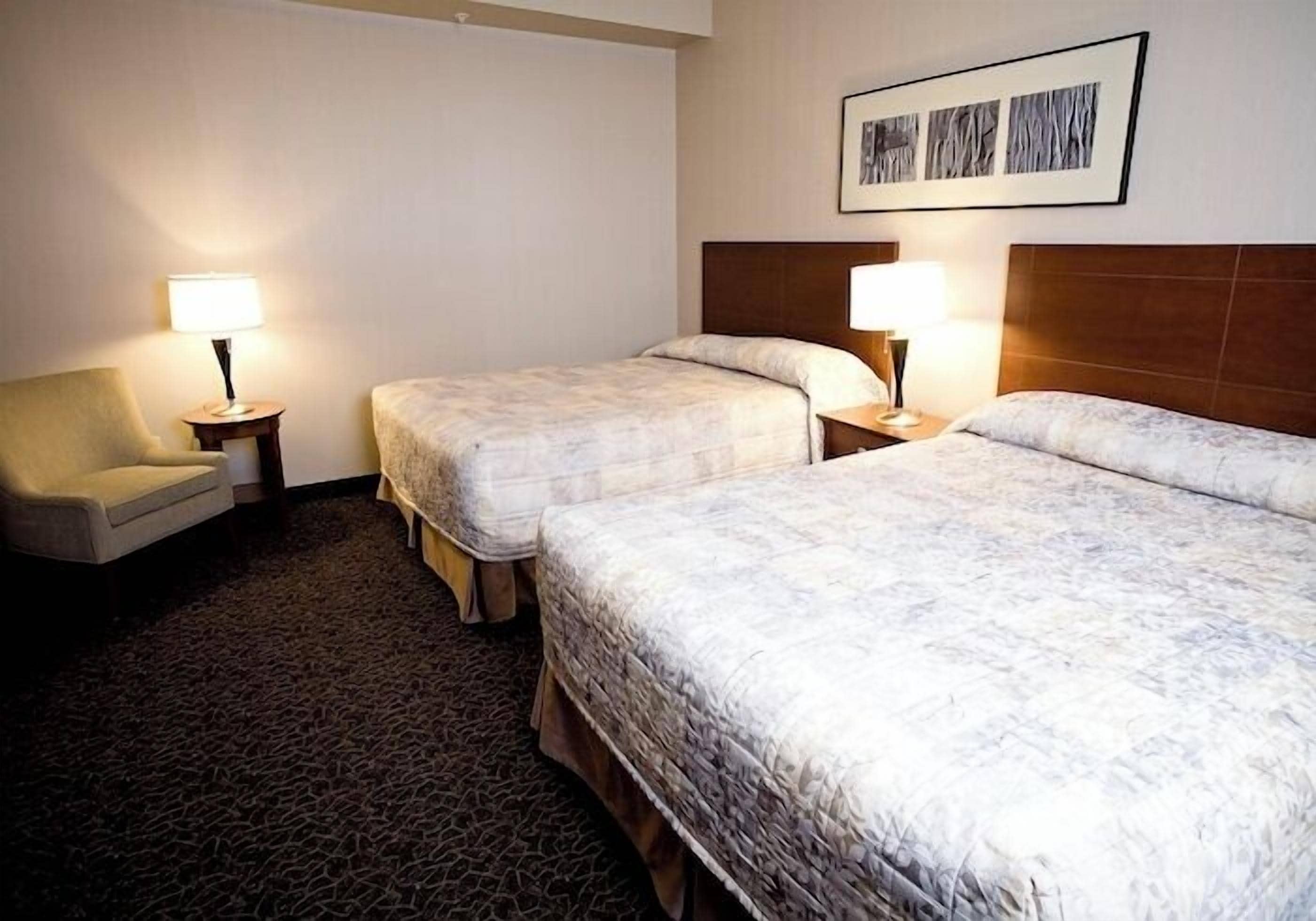 A hotel room with two beds, a chair, and a small table with lamps.