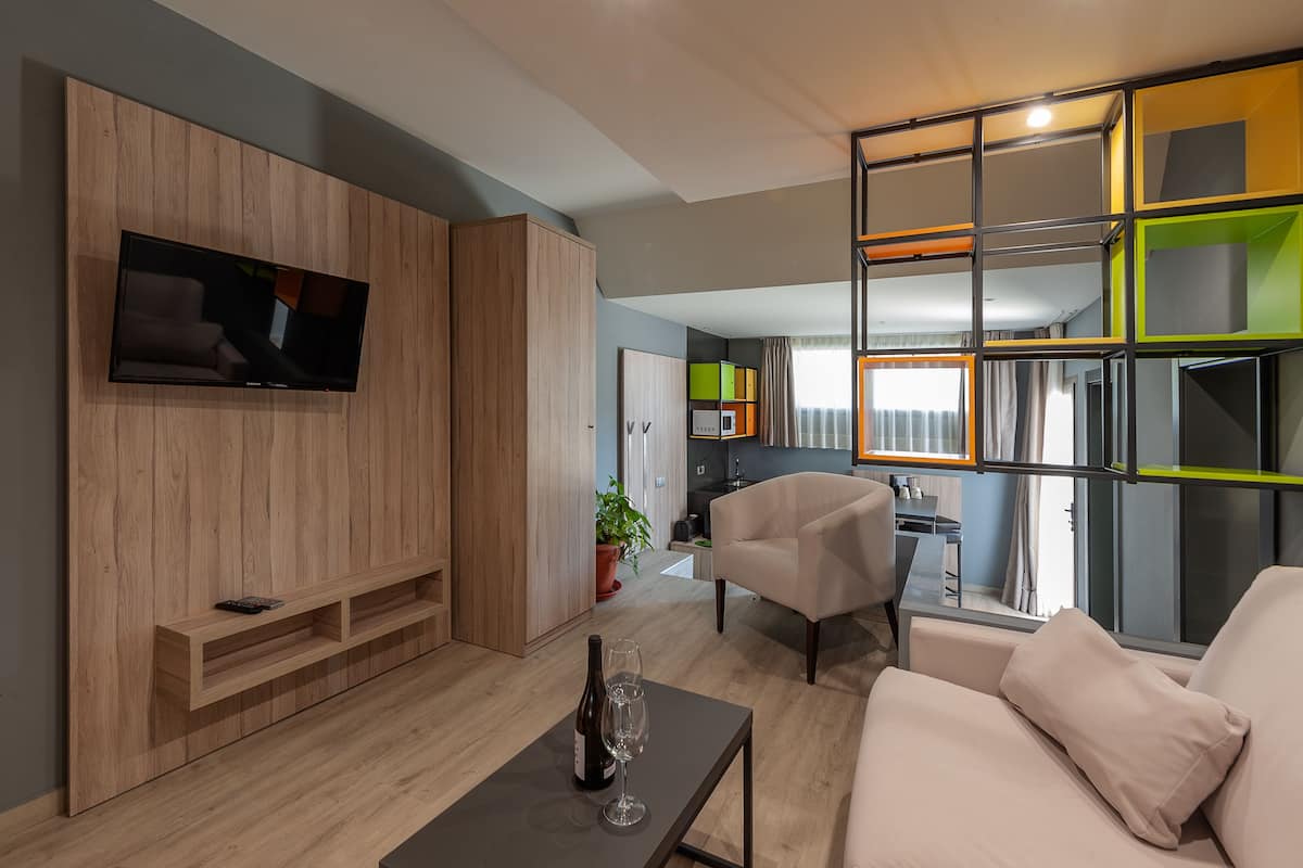 A modern living room with a flat-screen TV, a sofa, a coffee table with a bottle and glass, and a dining area in the background.