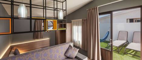Een moderne hotelkamer met een bank, een hanglamp en een schuifpui naar een balkon met buitenmeubels.
