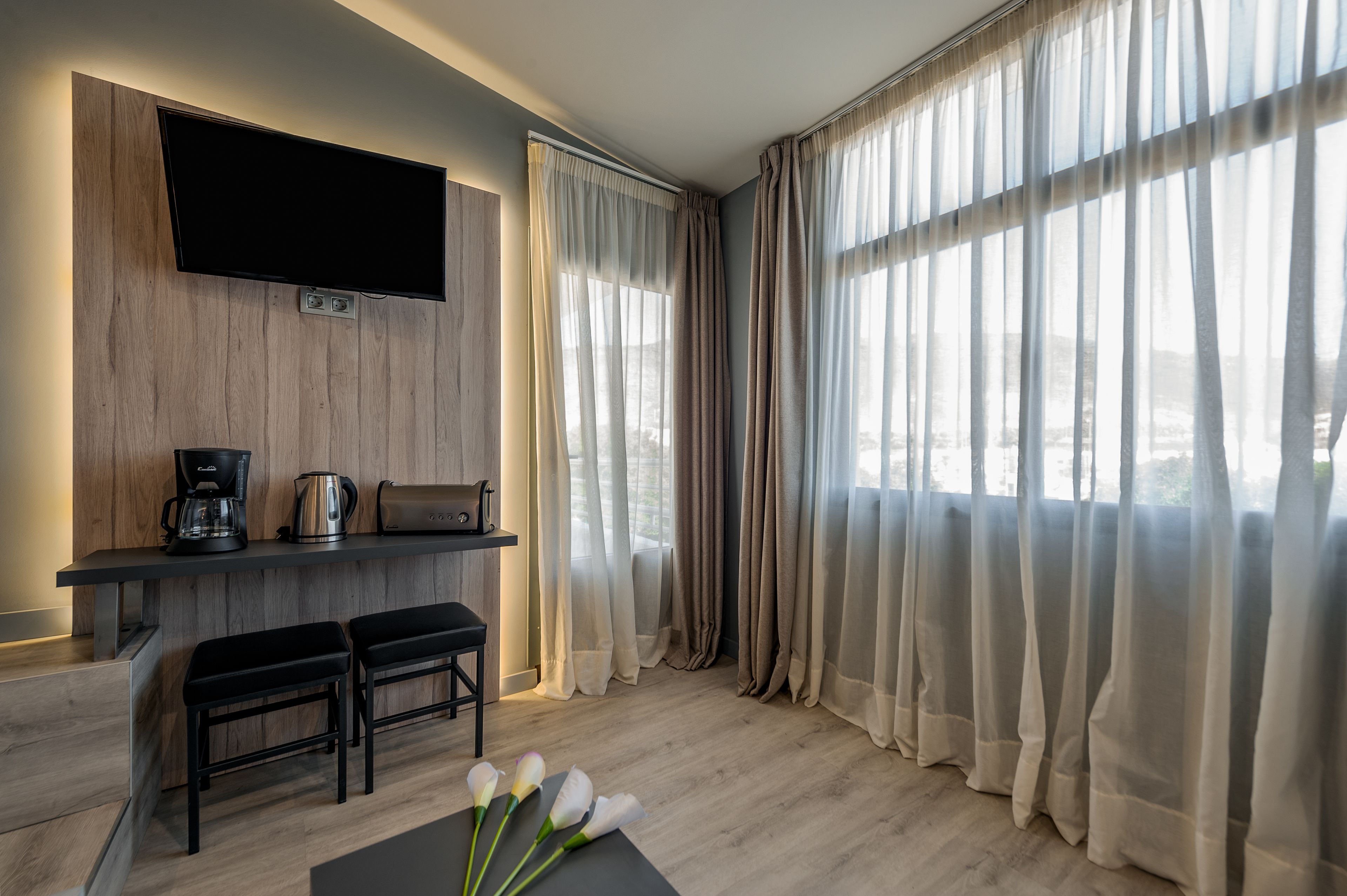 A modern room with a flat-screen TV mounted on the wall, a coffee station, two black bar stools, and large windows with sheer curtains.