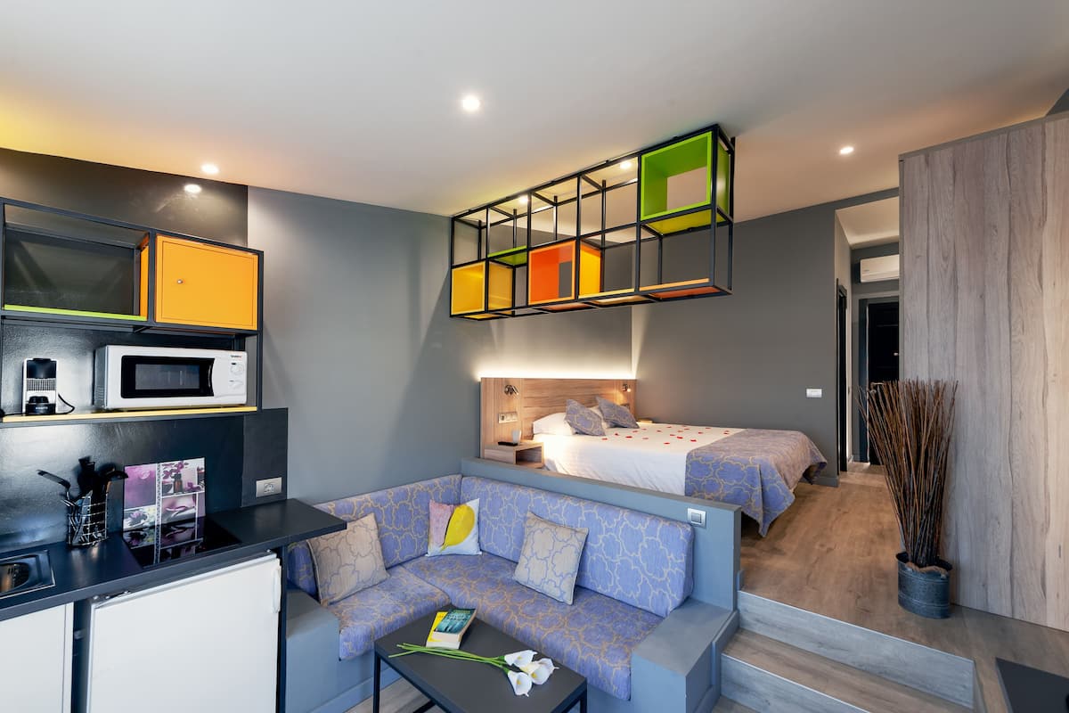 A compact hotel room with a kitchenette, a sofa, a bed, and a unique ceiling light fixture.