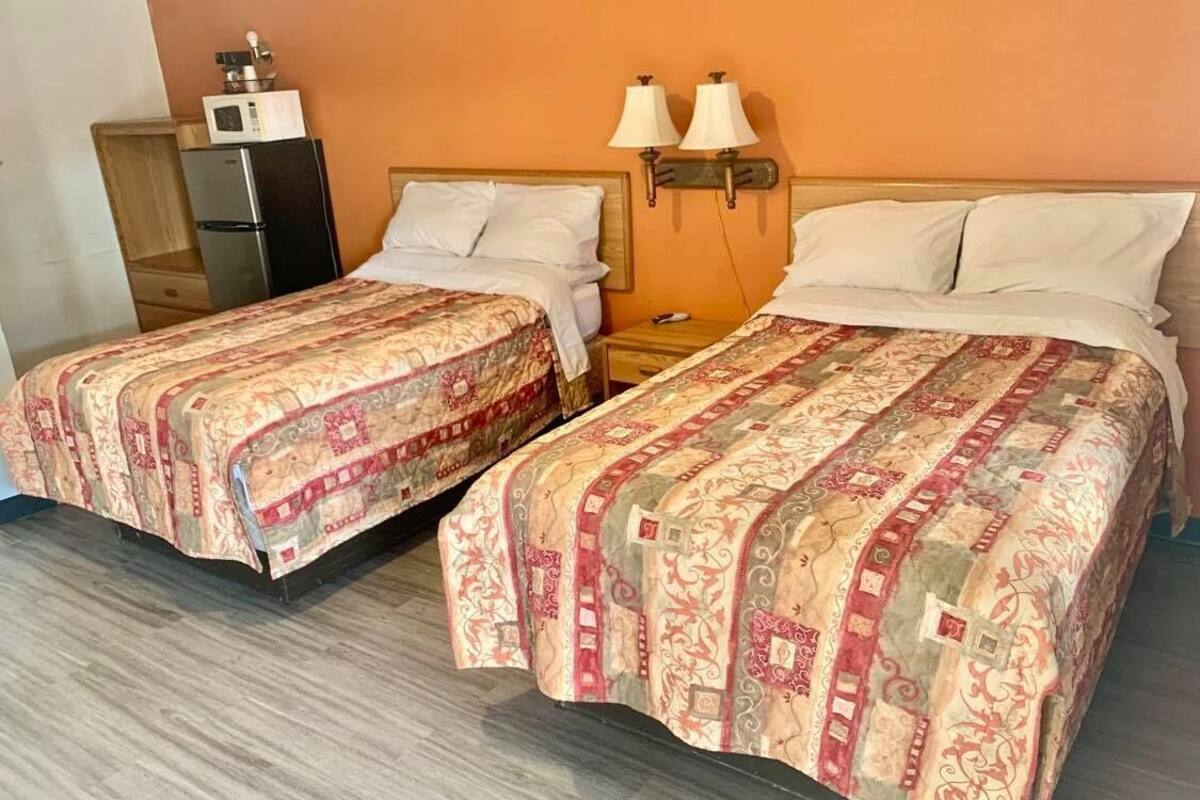 A hotel room with two beds, each with a patterned bedspread, a wooden nightstand, a wall-mounted lamp, and a refrigerator.