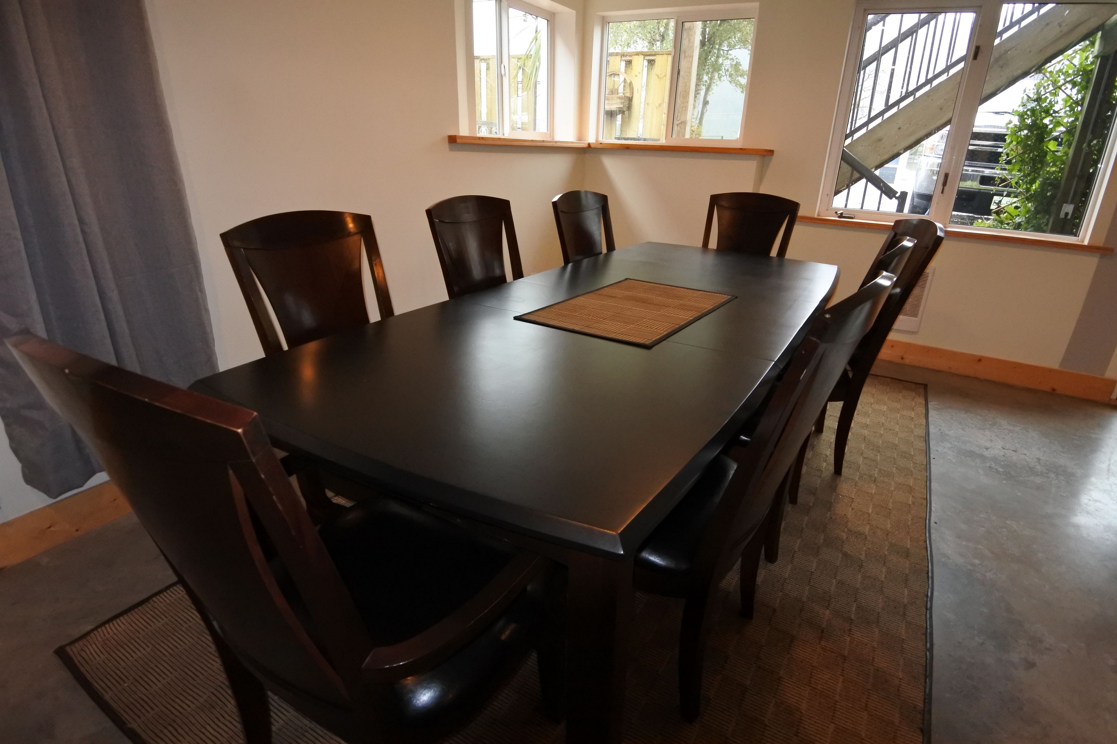 Un coin repas comprenant une table rectangulaire, six chaises et une fenêtre donnant sur un bâtiment et de la verdure.