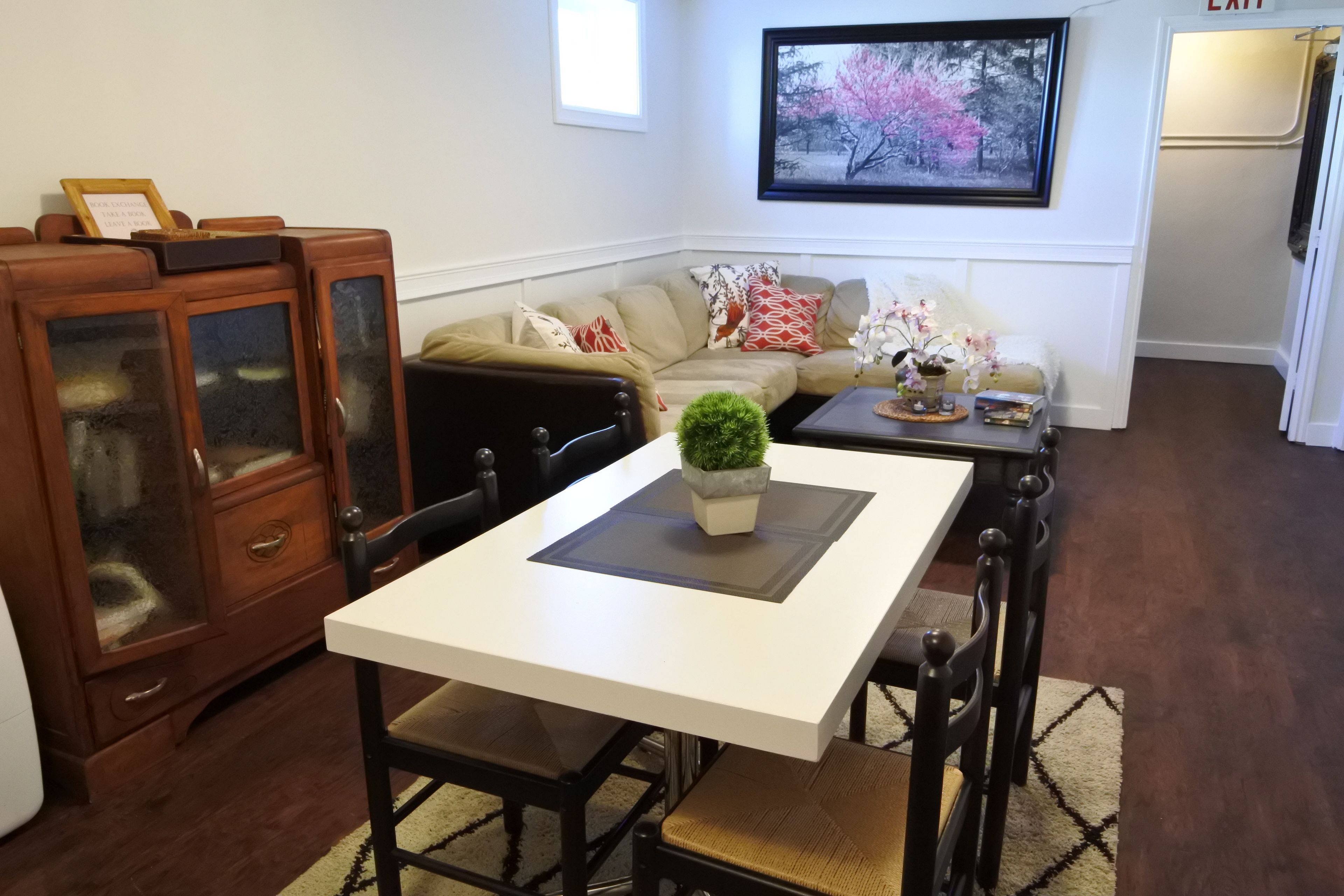 Een woonkamer met een witte tafel, donkerhouten stoelen, een beige bank met gekleurde kussens, een houten kast met glazen deuren, en een ingelijst schilderij aan de muur.