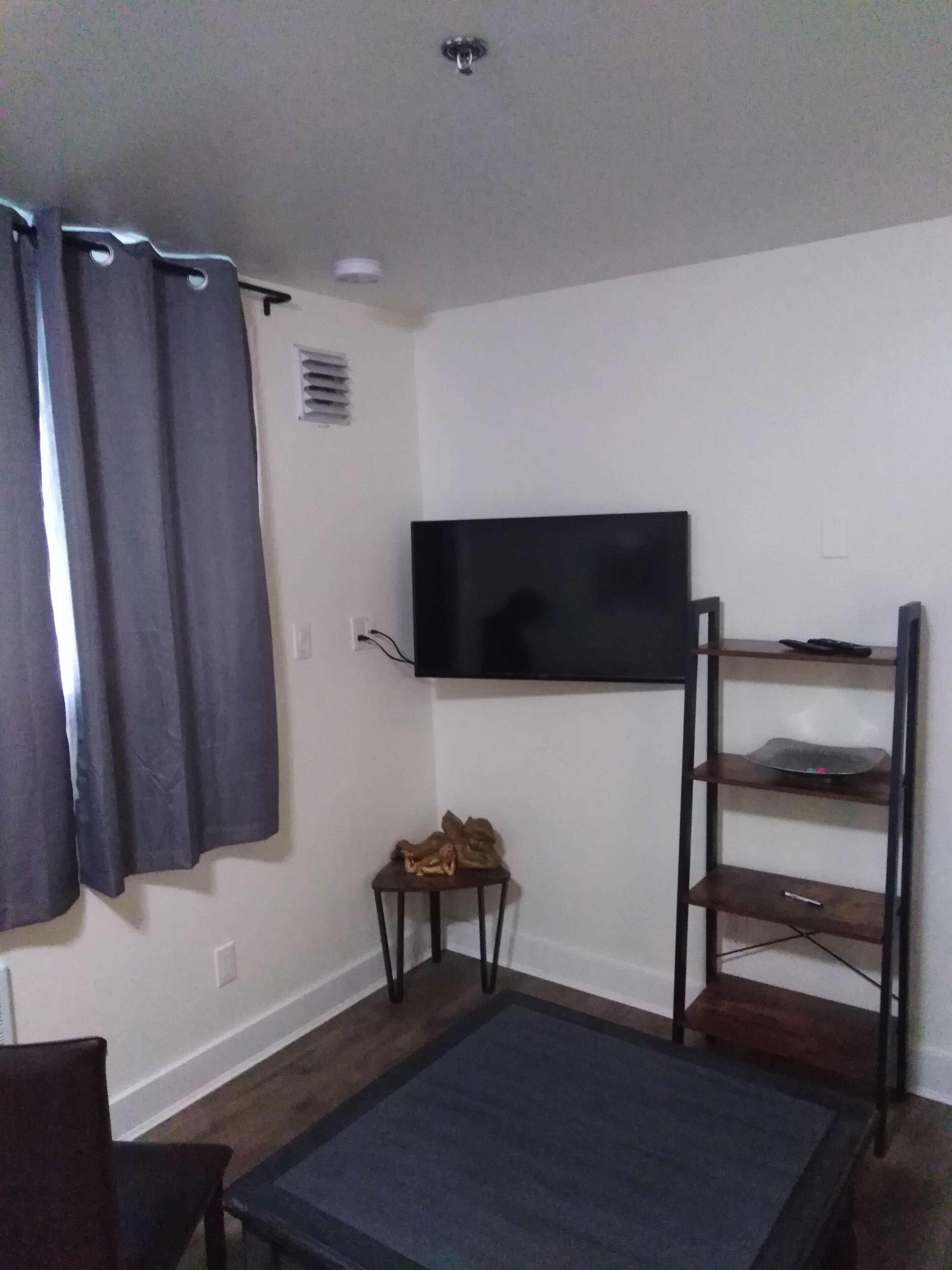 Una habitación pequeña y poco amueblada con una televisión de pantalla plana en la pared, una mesita de madera, una alfombra oscura y una silla.